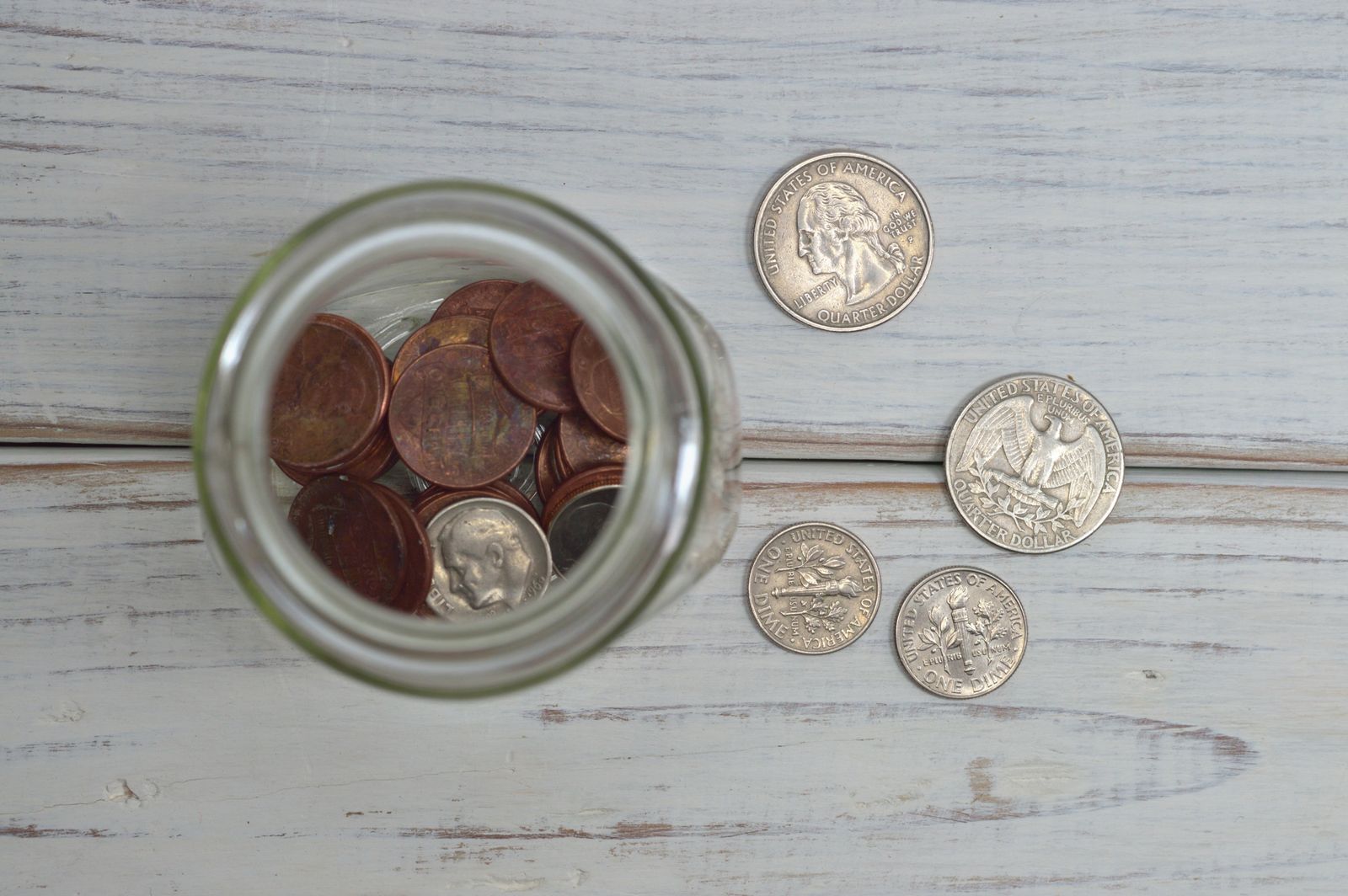 Coins in a glass jar next to loose change on a weathered wooden table