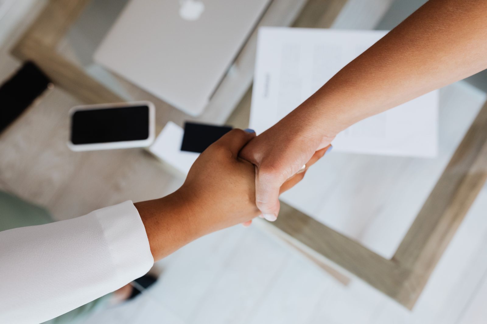Two people shaking hands over a desk with a laptop and paperwork